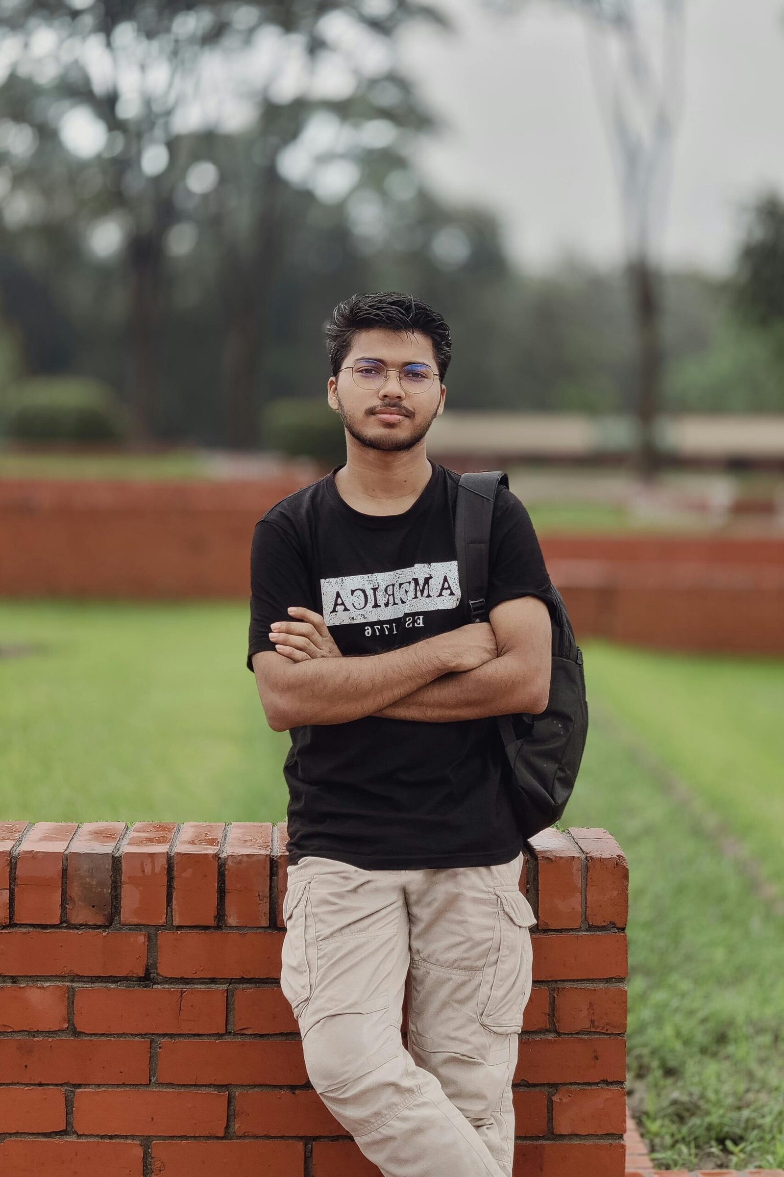 Young man with glasses in casual attire, standing confidently in a garden setting.