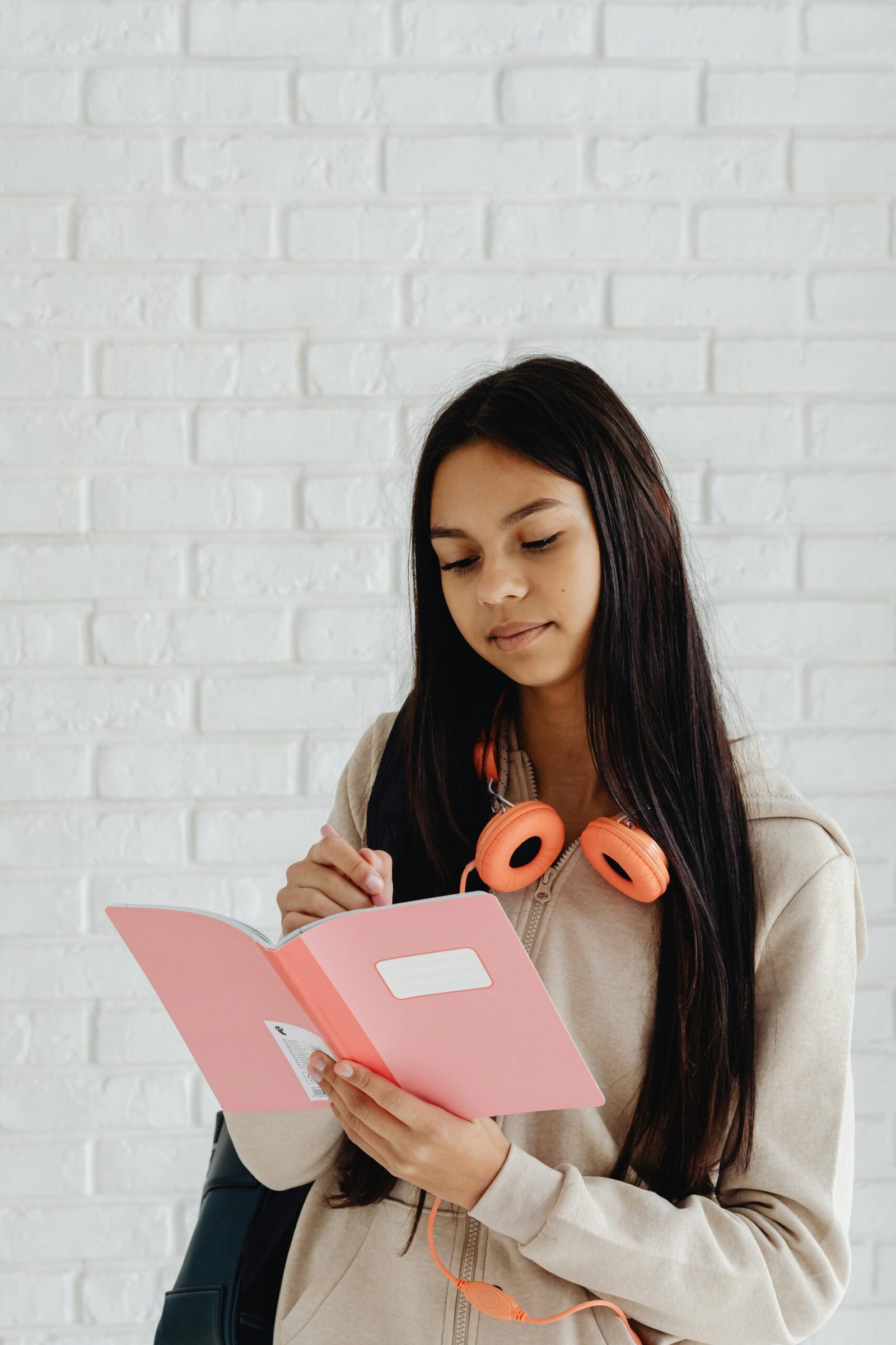 Teenager with long hair writes in a pink notebook, wearing headphones.