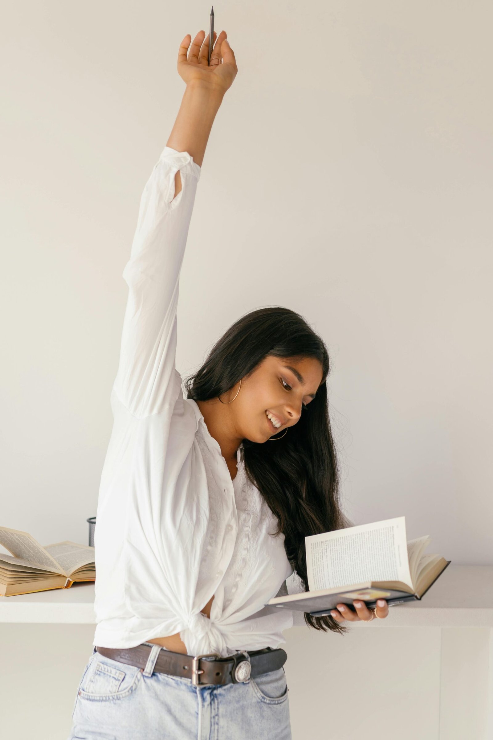 Smiling young woman indoors, raising her hand in celebration while studying a book.